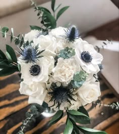 a bouquet of white and blue flowers sitting on top of a wooden table with greenery