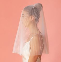 a woman wearing a white veil with sheer fabric on her head and shoulders, standing in front of a pink background