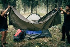 two people standing next to a tent in the woods with one person holding it up