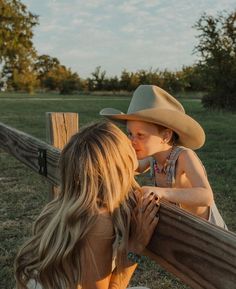 a woman and child leaning against a wooden fence in front of a field with trees