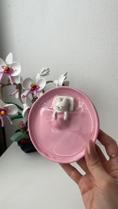 a hand holding a pink plate with a small white elephant on the bottom and flowers in the background