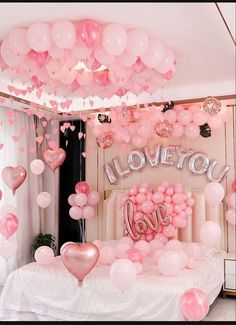 a bed topped with lots of pink balloons and heart shaped balloons hanging from the ceiling