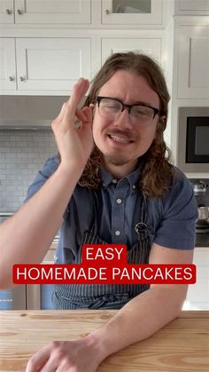 a man with long hair and glasses sitting at a table in front of a wooden cutting board