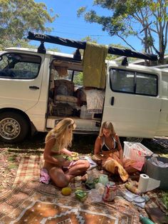 two women sitting on the ground in front of a van eating food and talking to each other