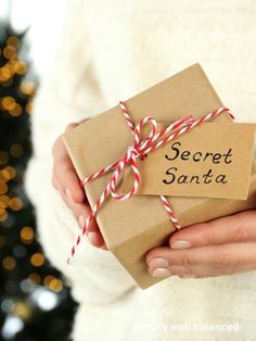 a woman holding a wrapped present box with the words secret santa written on it in front of a christmas tree