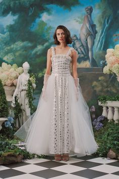 a woman in a white dress standing on a checkered floor with flowers and statues behind her