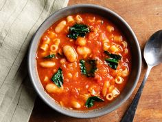 a bowl filled with pasta and spinach on top of a wooden table next to a spoon