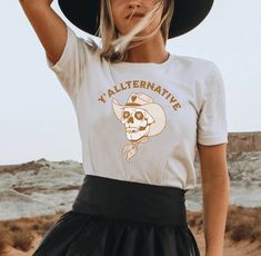 Introducing our exclusive Yallternative Shirt, a perfect blend of southern charm and alternative style! Embrace your inner emo country enthusiast with this eye-catching Yallternative T-Shirt featuring a mesmerizing cowgirl skull design. With its gothic cowgirl aesthetic and a touch of Texas goth, this Alt Girl Shirt is an absolute must-have for anyone seeking a unique and edgy fashion statement. Unleash your yallternative spirit and proudly show off your love for the alt country scene with this captivating and rebelliously chic garment. Get ready to turn heads and set trends with this extraordinary Alt Country Shirt! Overview -These t-shirts have-ribbed knit collars to bolster shaping. -The shoulders have taping for better fit over time. -Dual side seams hold the garment's shape for longer White Tops For Alternative Fashion In Fall, Fall Graphic Tee For Alternative Fashion, Goth Country, Gothic Cowgirl, Emo Cowgirl, Alt Country, Western Gothic, Cowgirl Aesthetic, Alternative Style