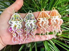 "These Jellyfish earrings are made of resin and various glitters. Each one is unique and one of a kind, due to the nature of resin. The etched design is hand painted. These would be a lovely accessory in the summer, at the beach or a trip to the aquarium.  The orange option has a slight imperfection on the back of one of the earrings. Nothing wrong with it and barely noticeable, just a little extra resin got on it so there's a couple smooth bumps which is why we're lowering the price of that pair slightly.   These items are handmade and hand painted in our living room. They take around an hour to finish painting. We are not a big shop or corporation, just a couple pursuing their passion for creation. With the nature of handmade items like this, there may be some tiny imperfections we misse White Resin Earrings For Summer, Resin Earrings For Summer Beach, Summer Beach Resin Earrings, White Resin Earrings For The Beach, Jellyfish Earrings, Summer At The Beach, The Aquarium, North Hollywood, Etsy Earrings Dangle