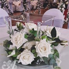 white roses and greenery are arranged in a square vase on a table at a wedding reception