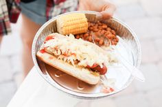 a person holding a hot dog and corn on the cob in a tin container