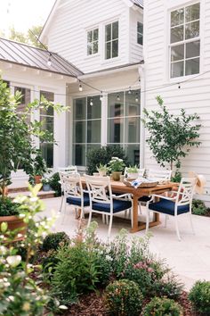 an outdoor dining table and chairs in front of a white house with potted plants