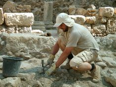 a man kneeling down next to a bucket filled with dirt and rocks while holding a shovel