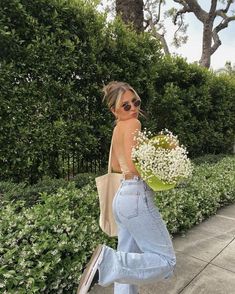 a woman walking down the street with flowers in her hand and wearing high rise jeans