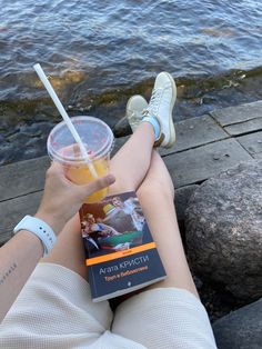 a person sitting on the ground with a book and drink in their hand next to water