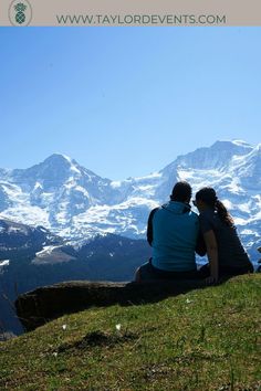 two people sitting on top of a mountain looking at the snow covered mountains in the distance