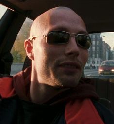 a bald man wearing sunglasses sitting in the back seat of a car on a city street