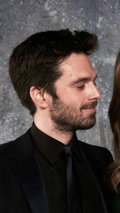 a man and woman standing next to each other in front of a stone wall wearing black suits