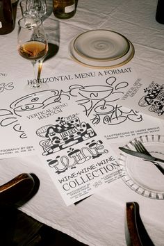 a table topped with wine glasses and menus next to plates on top of a white table cloth