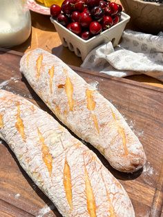 two loaves of bread sitting on top of a wooden cutting board next to bowl of cherries