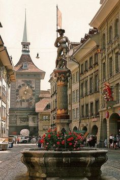 a fountain in the middle of a street with flowers on it and a clock tower in the background