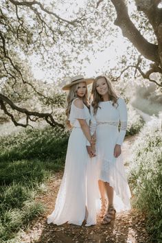 two women in white dresses and hats posing for a photo under the shade of trees