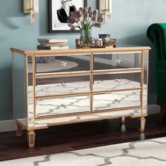 a gold and mirrored dresser with flowers on top in front of a green chair next to a blue wall