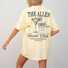 a woman wearing a yellow t - shirt that says the allen social club and has a cocktail in it