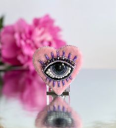 a pink heart shaped brooch with an eye on it's side and flowers in the background