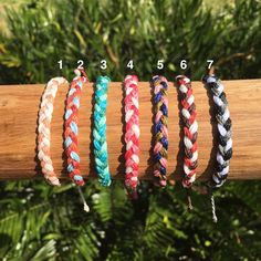 five different braided bracelets sitting on top of a wooden stick in front of some trees