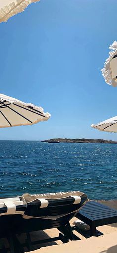 two beach chairs with umbrellas over them on the sand near the water and an island in the distance