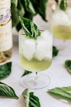 two glasses filled with ice and green leaves on top of a white table next to bottles