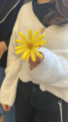 a woman holding a yellow flower in her right hand while wearing a white sweater and black pants