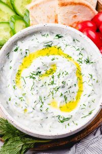 a white bowl filled with dip surrounded by sliced cucumbers, tomatoes and bread