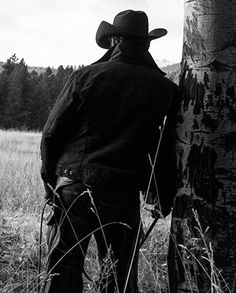 a man wearing a cowboy hat standing next to a tall tree in a grassy field