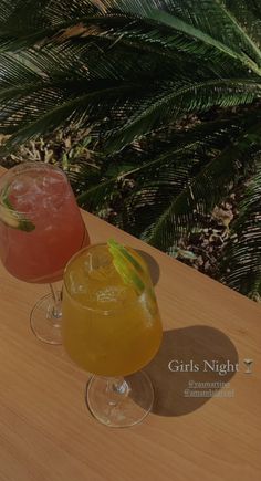 two cocktails sitting on top of a wooden table next to a palm leaf covered tree