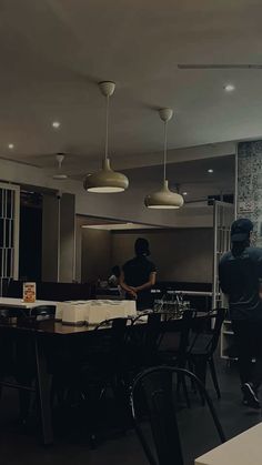 two people standing in a room with tables, chairs and lamps hanging from the ceiling