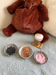 a brown teddy bear sitting on top of a bed next to two bowls of food