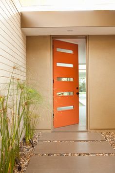 an orange door is in front of a white wall and some green plants on the floor