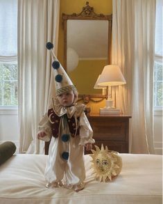 a baby dressed up as a jester standing on top of a bed next to a stuffed animal