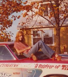 a woman sitting on the hood of a race car in front of a house and tree