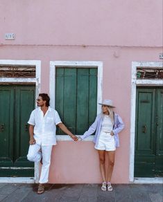 a man and woman holding hands while standing in front of a pink building with green doors