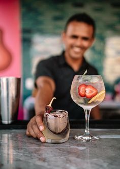 a man behind a bar holding a glass with a strawberries in it and another drink on the counter