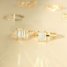 Two diamond rings shine on a flat surface. The stones, so vibrant, the rings refract little rainbows onto the table. Baguette Cut Moissanite Solitaire Ring, Moissanite Solitaire Ring In Baguette Cut, Moissanite Single Diamond Wedding Ring, Minimalist Moissanite Emerald Cut Ring, Moissanite Fine Jewelry With Single Diamond, Minimalist Emerald Cut Moissanite Ring, Radiant Cut Single Diamond Moissanite Wedding Ring, Classic Baguette Cut Single Diamond Ring, Solitaire Moissanite Diamond Ring Radiant Cut