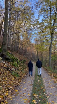 two people walking down a dirt road in the woods with leaves on the ground and trees all around them