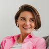 a smiling woman sitting on top of a couch next to a gray pillow and pink shirt