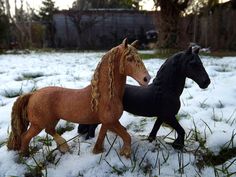 two toy horses standing in the snow on top of grass and bushes, one black and one brown