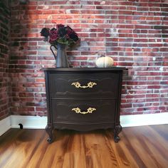 a black dresser with gold trim and flowers in a vase on top, against a brick wall