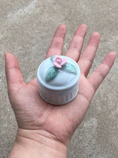 a hand holding a small white container with a pink flower on it's lid