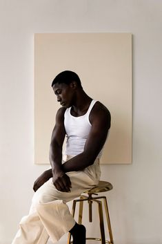 a man sitting on top of a wooden stool next to a white wall with a painting behind him